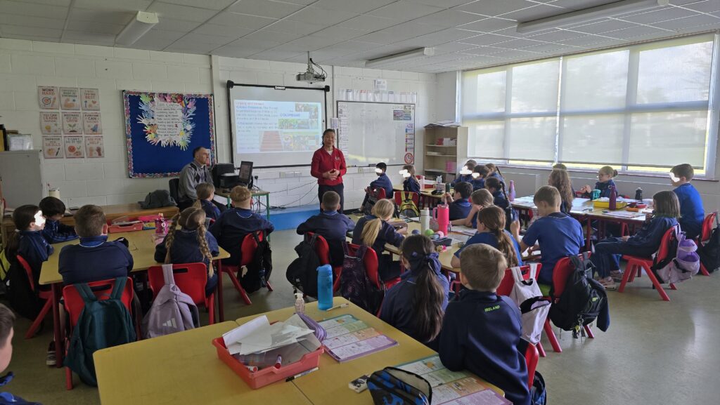 Teacher stands at the front of a classroom presenting to a group of students at desks.