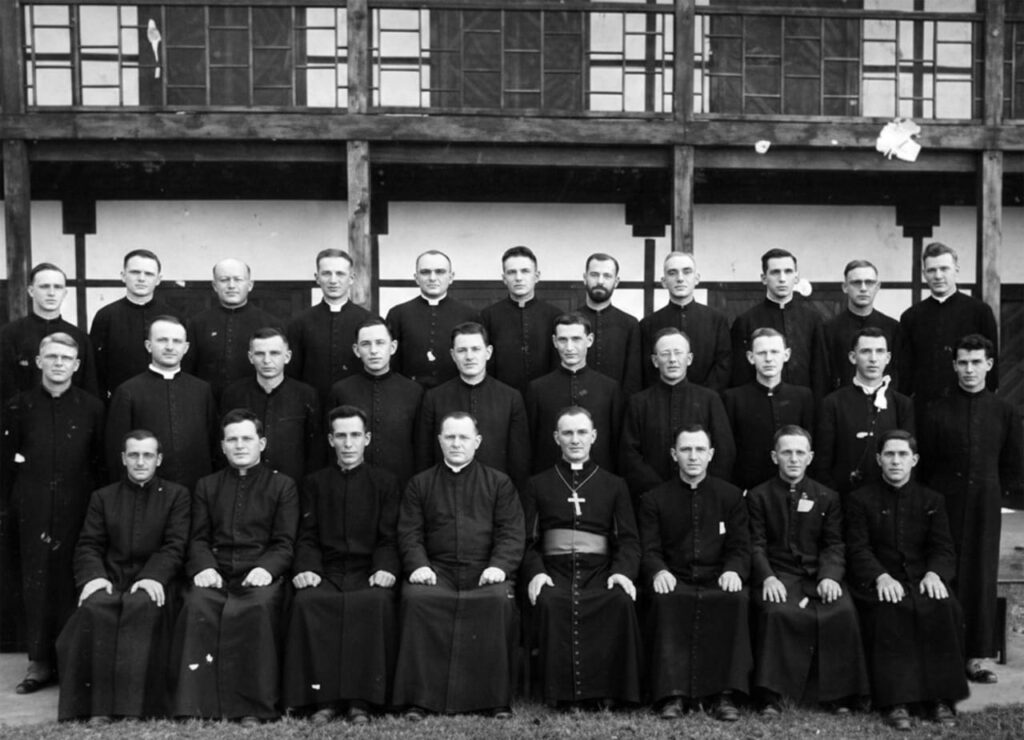 Group of Catholic priests in black cassocks posing for a formal photo outside a wooden building.
