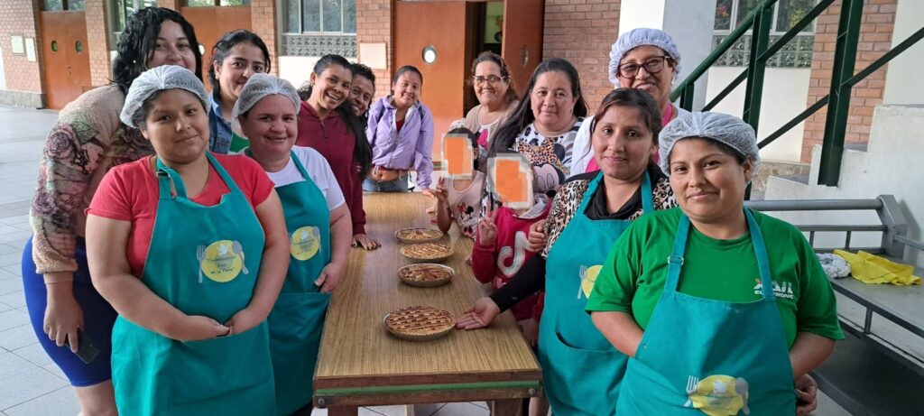 Group of women in teal aprons and hairnets stand around a table with several pies, smiling in a courtyard.