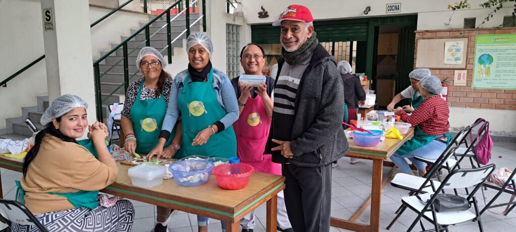 02 - Columban Missionaries Group of volunteers wearing hairnets and aprons stand around a table preparing food at a community kitchen.