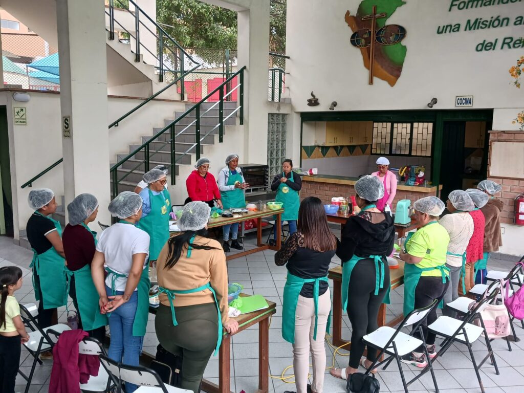 03 - Columban Missionaries Group of people in teal aprons and hair nets watching a cooking demonstration at a community kitchen center, outdoors.