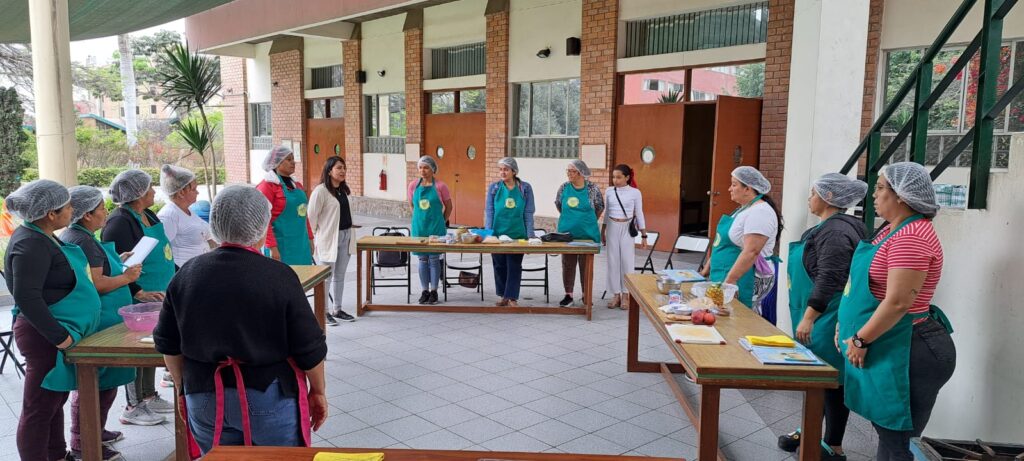 04 - Columban Missionaries Group of people in aprons and hairnets stand in a semicircle around kitchen tables with ingredients, in a covered outdoor area (cooking class).