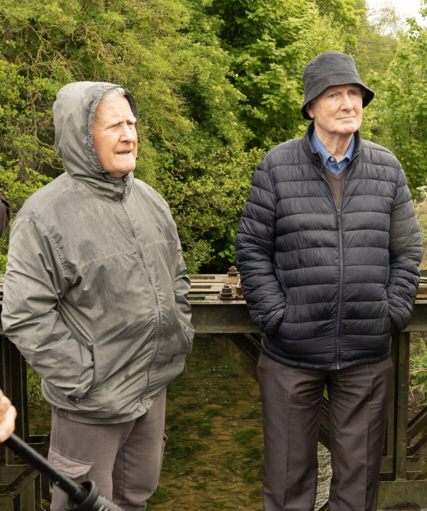 10 Bring me to the River - Columban Missionaries Two elderly men in jackets stand on a wooden railing outdoors, with lush green trees behind them.