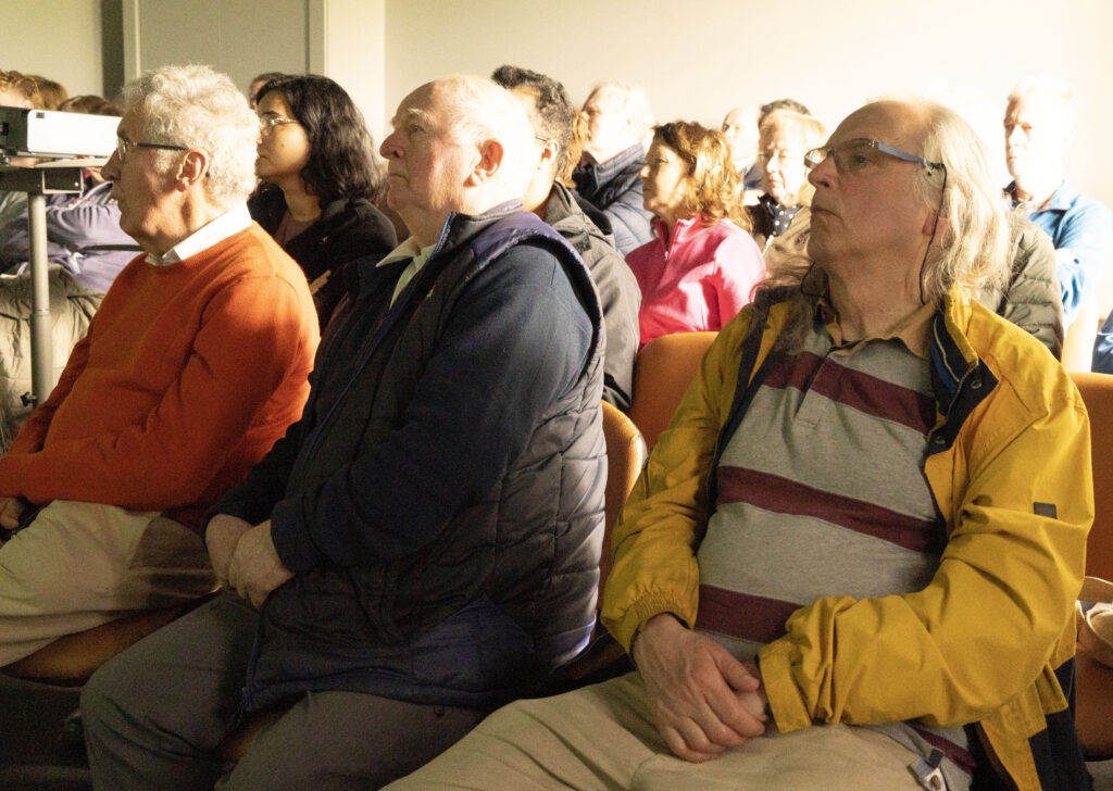 5 Bring me to the River - Columban Missionaries Audience of adults seated in a conference room wearing jackets, facing toward the front; projector visible on the left side.