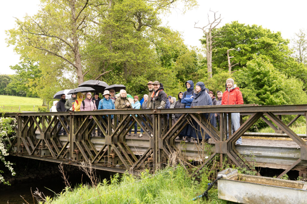 8 Bring me to the River - Columban Missionaries Group of people standing on a wooden bridge in a park, many holding umbrellas in the rain with green trees in the background.