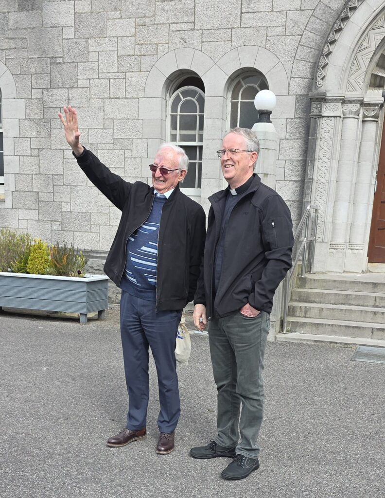 Luxeuil Pilgrims10 - Columban Missionaries Two older men stand outside a stone building; one waves with his right hand while the other smiles nearby.