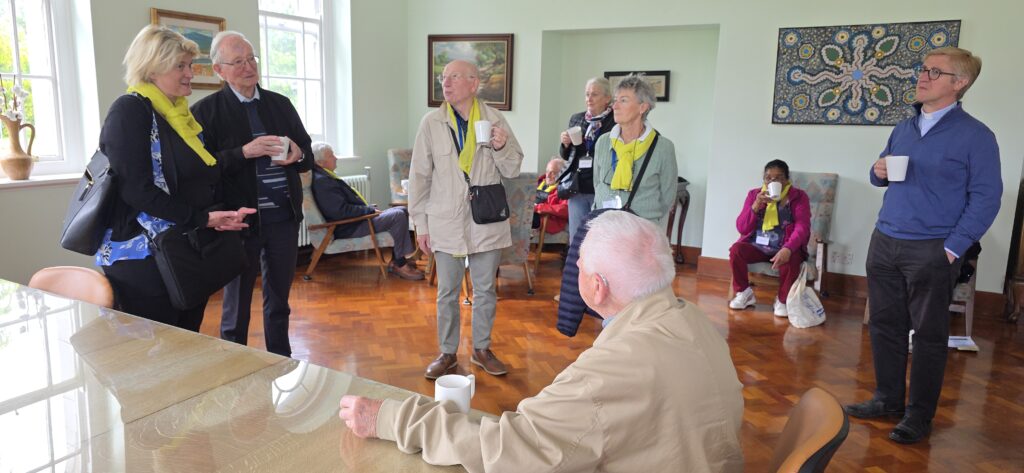 Luxeuil Pilgrims4 - Columban Missionaries Group of senior adults standing and chatting in a bright lounge, each holding a mug, some wearing yellow scarves and name badges.