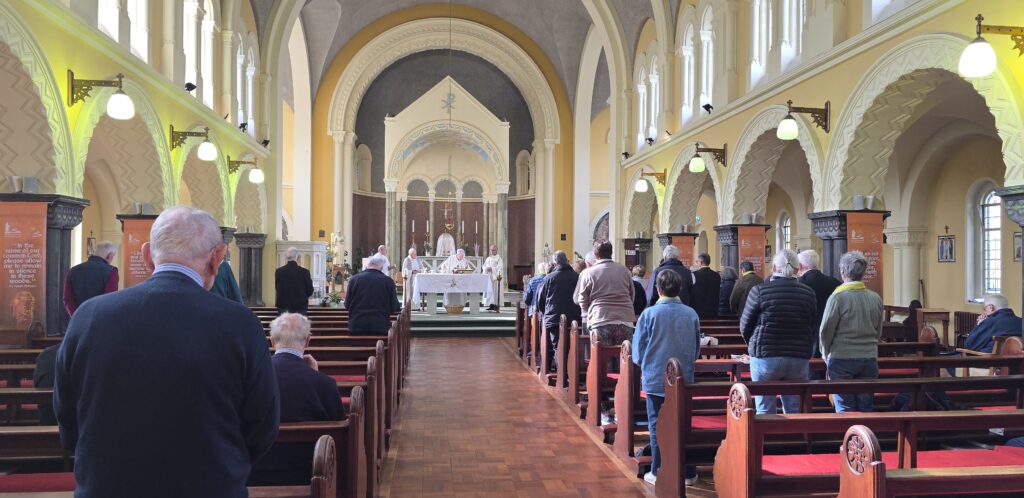 Luxeuil Pilgrims6 - Columban Missionaries Interior of a parish church with tall arches, yellow walls, and a congregation standing in wooden pews during a service at the altar area.