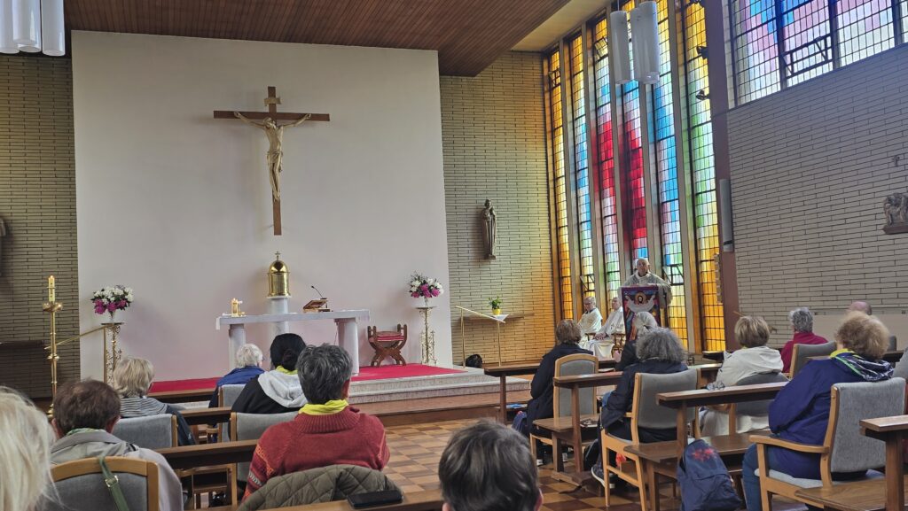 Luxeuil Pilgrims7 - Columban Missionaries Church sanctuary with a large crucifix over the altar, candles and flower arrangements, and worshippers seated as a priest speaks near the stained glass windows on the right.