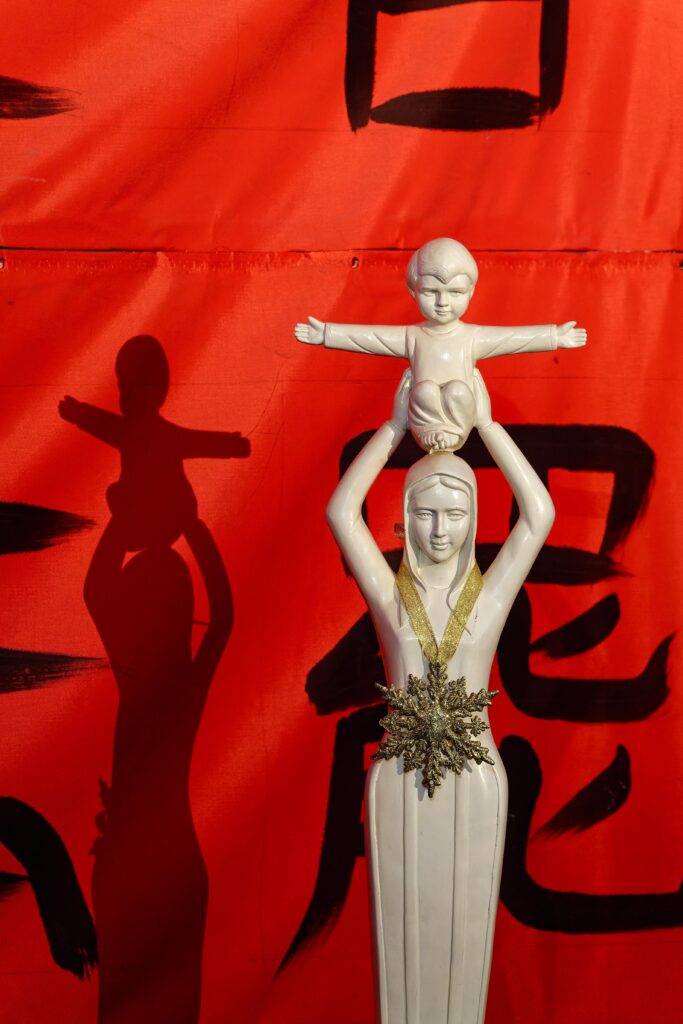 White statue of a person holding a child on their shoulders, with a gold sash and a snowflake ornament, against a bright red wall with a shadow to the left.
