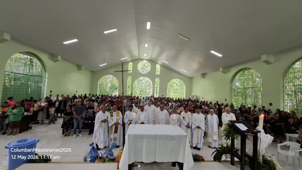 Inside a church during a Mass, priests in white vestments stand at the altar as a large congregation fills the pews.