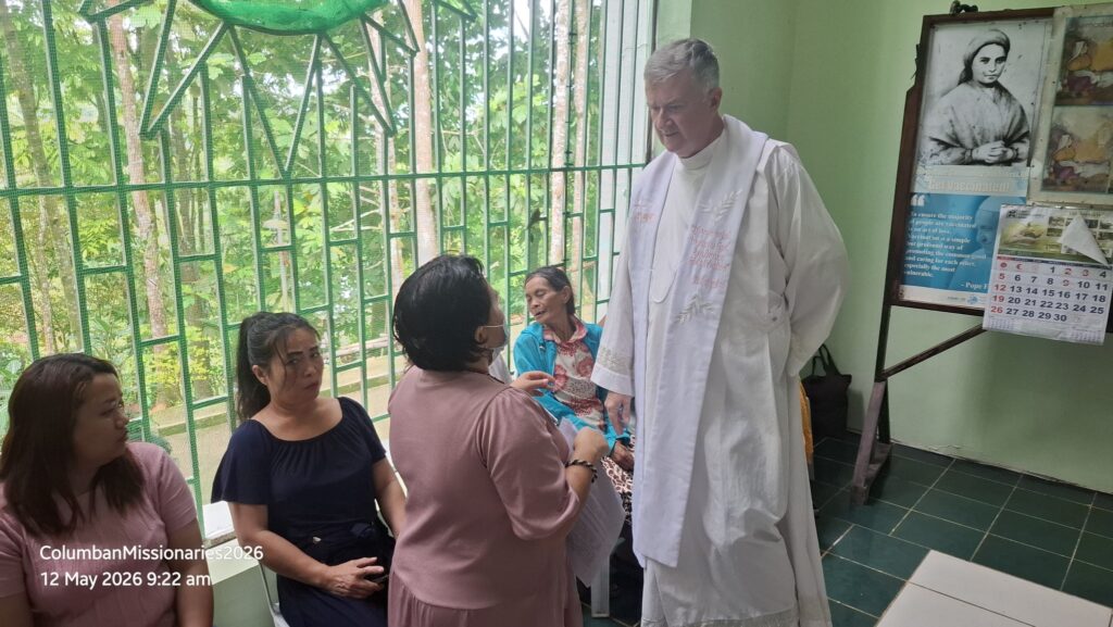 Priest in white vestments speaking with seated women in a bright room with green window bars and a wall calendar/posters nearby.