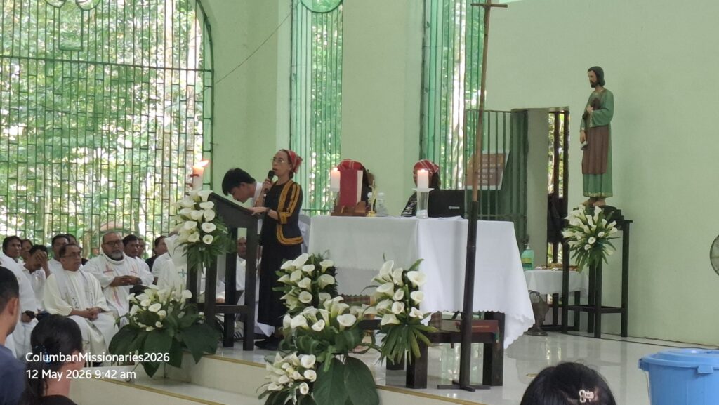 Woman wearing a headscarf speaks into a microphone at a lectern during a church service, with clergy in white robes seated nearby and white floral arrangements on the altar aisle.