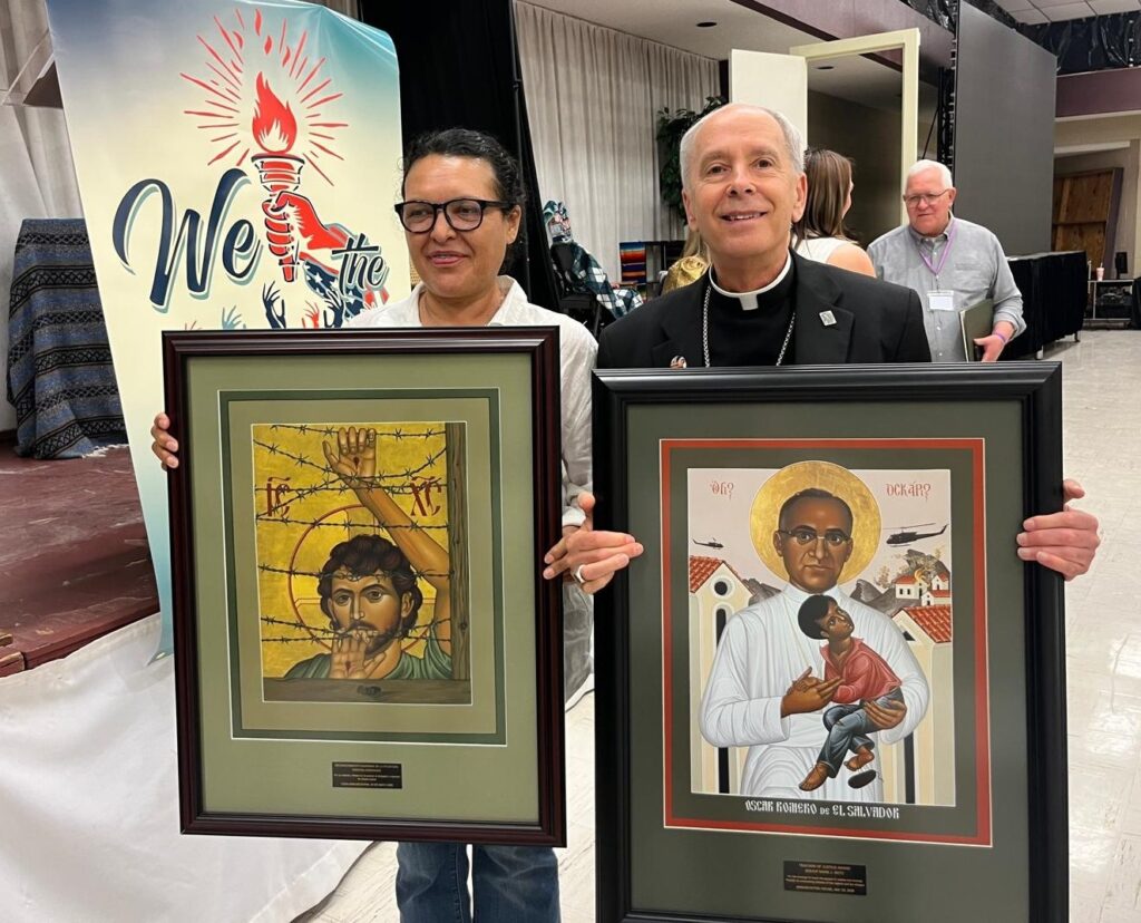 Two people, a woman and a priest, hold large framed religious icons at an event, with a banner in the background reading 'We the'.