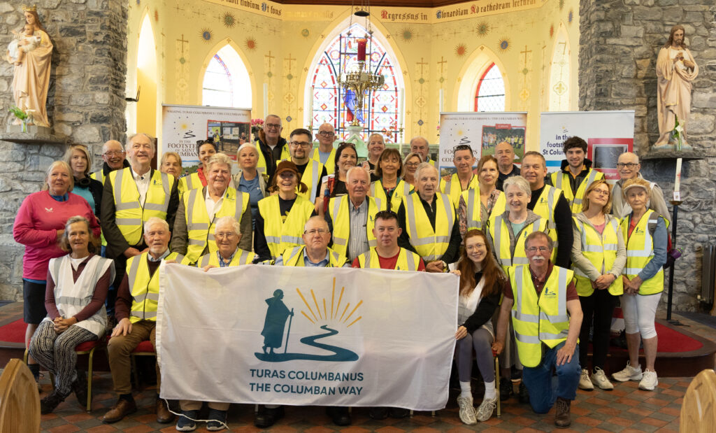 Group of volunteers in yellow safety vests pose inside a church with stained glass and statues, holding a banner for The Columban Way (Turas Columbanus).