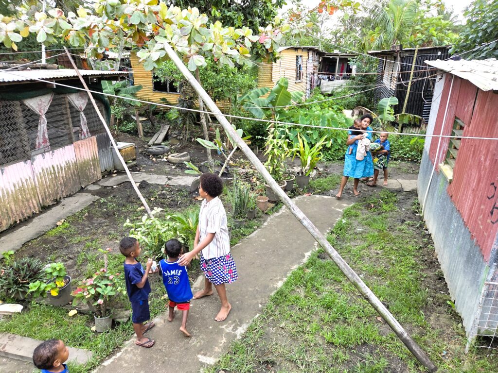 Children playing barefoot in a muddy, plant-filled yard between tin-shack houses with clotheslines overhead.