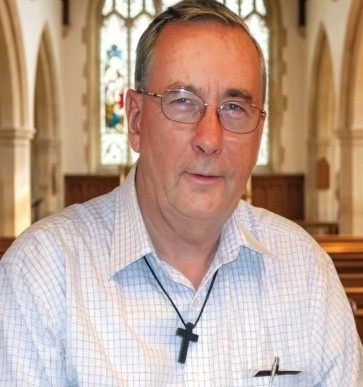 Fr Barney Martin A middle-aged man wearing glasses and a cross necklace sits inside a church pews with stained glass in the background.