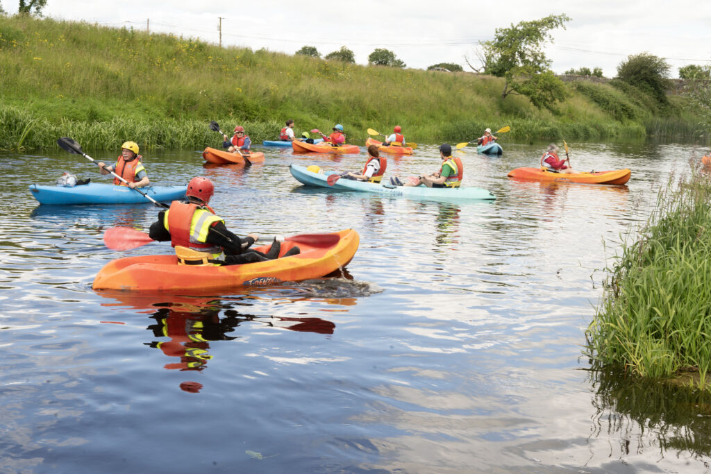 Pilgrims on the River Boyne - Columban Missionaries Group of kayakers in bright orange, blue, and teal boats paddling on a calm river beside a grassy embankment with trees in the background.