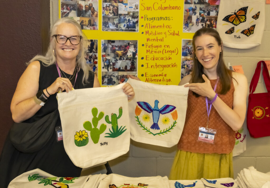 Two smiling women hold up handmade tote bags decorated with cactus and a blue bird at a community event display board behind them.