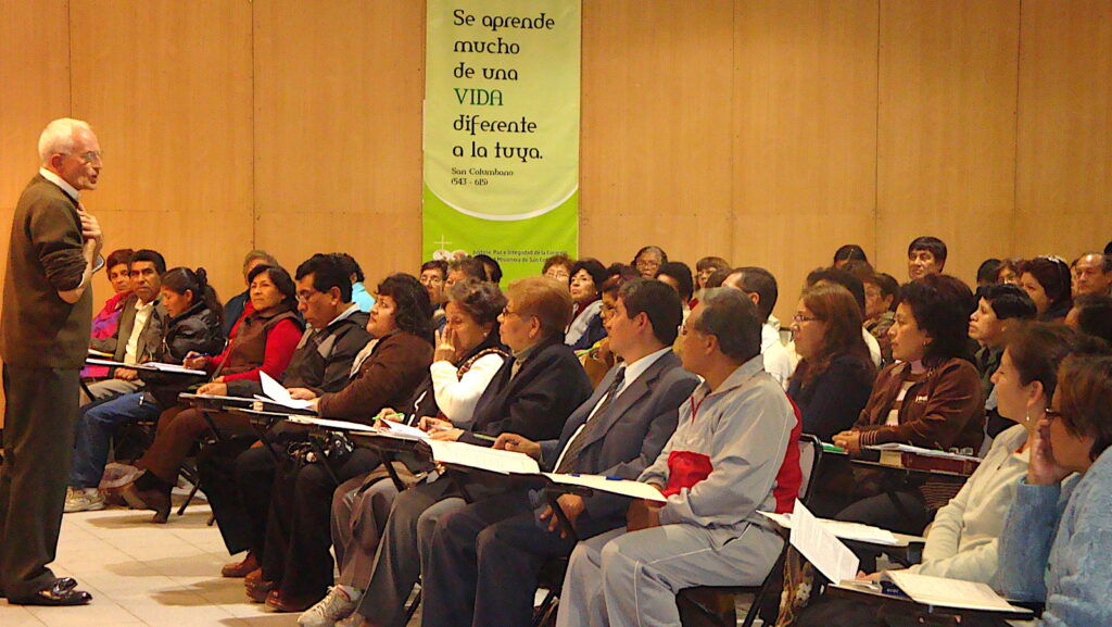 Older man standing at left speaking to a seated audience in a conference room; banner with Spanish quote in the background.
