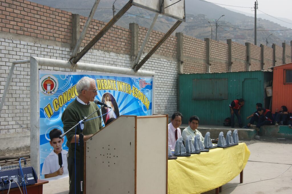 Older man speaks at a podium during an outdoor school debate event; banner and audience in background.