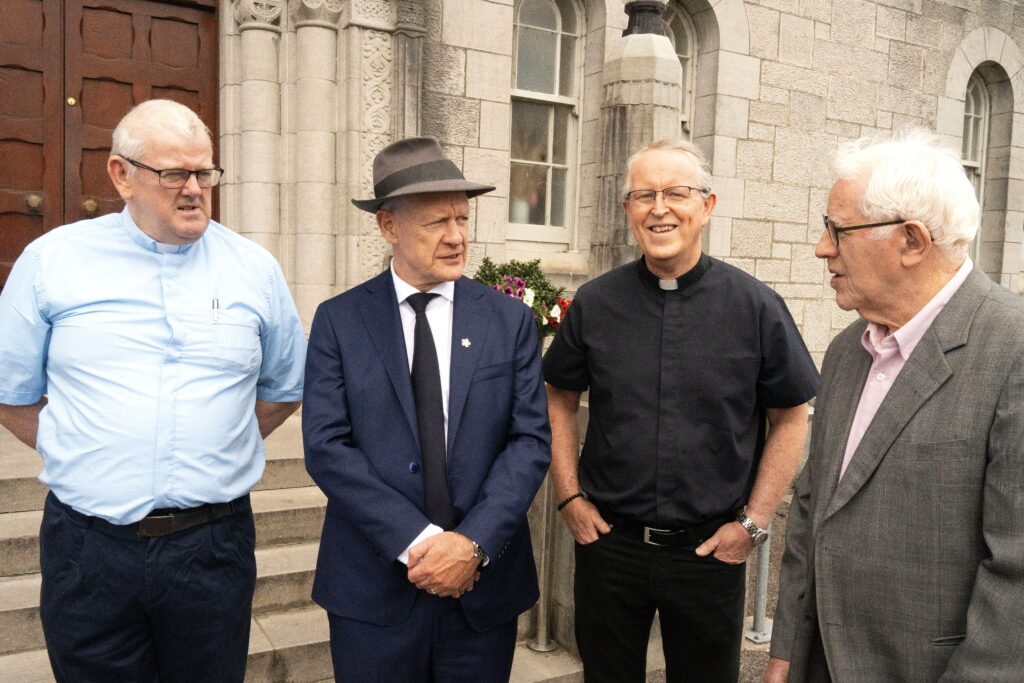 Four men standing outside a stone building, chatting; two wear clerical collars, one in a suit with a hat, and another in a light gray suit.