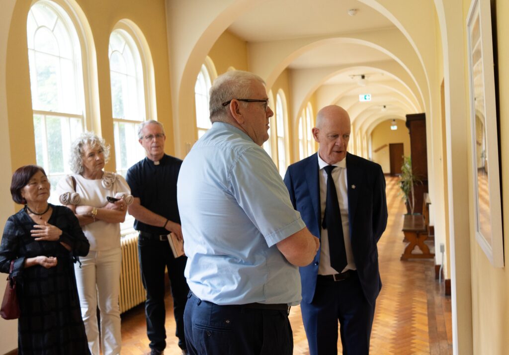 Group of adults standing in a sunlit arched hallway, chatting; one man in a blue shirt foreground, others in formal attire behind him
