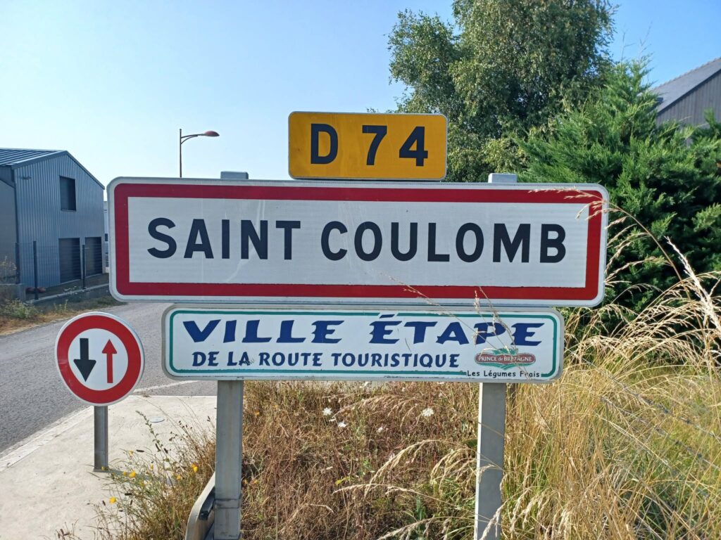 Road signs in Saint-Coulomb: a yellow D74 plate above a large white sign reading 'SAINT COLOMB', with a round red/white sign to the left.
