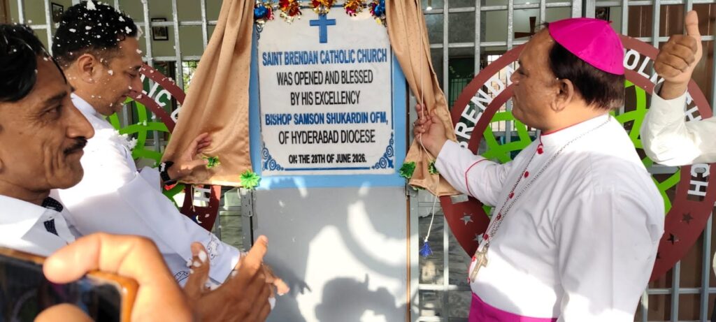 Bishop in white clerical robes and pink zucchetto unveils a sign announcing Saint Brendan Catholic Church opened and blessed by Bishop Samson Shukardin, Hyderabad Diocese, with two men in white shirts nearby.