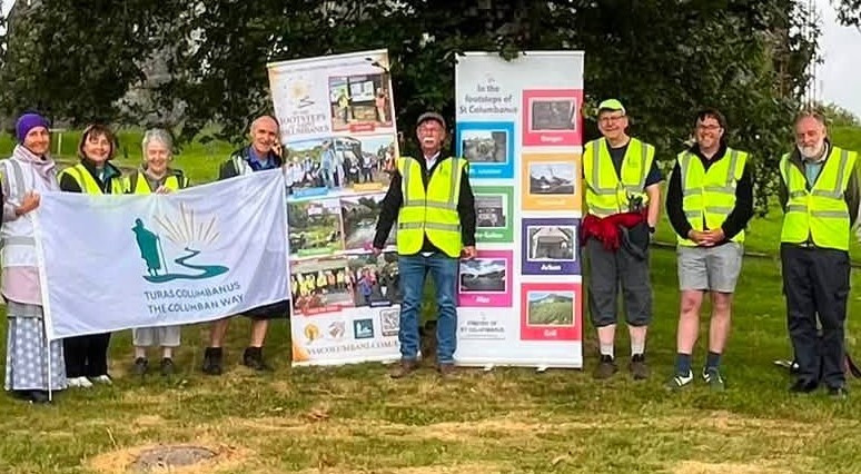 Nine volunteers in high-visibility vests stand on grass beside tall information banners and a white flag with a logo.