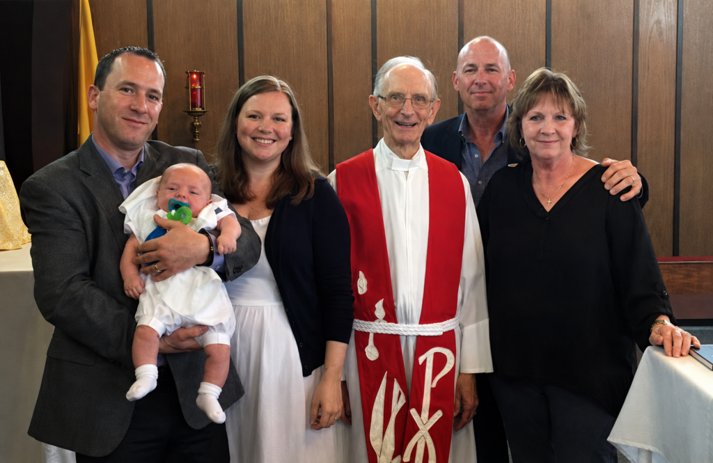 Father in a suit holding a baby, flanked by a smiling couple, a clergyman in red vestments, and a woman in black at a church event.