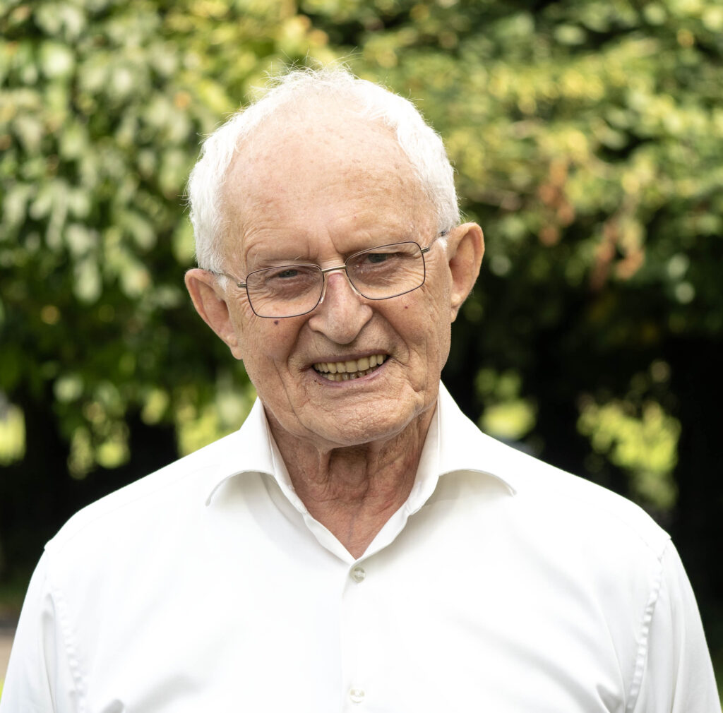 Close-up portrait of an elderly man with white hair and glasses outdoors, wearing a white shirt, smiling with greenery in the background.