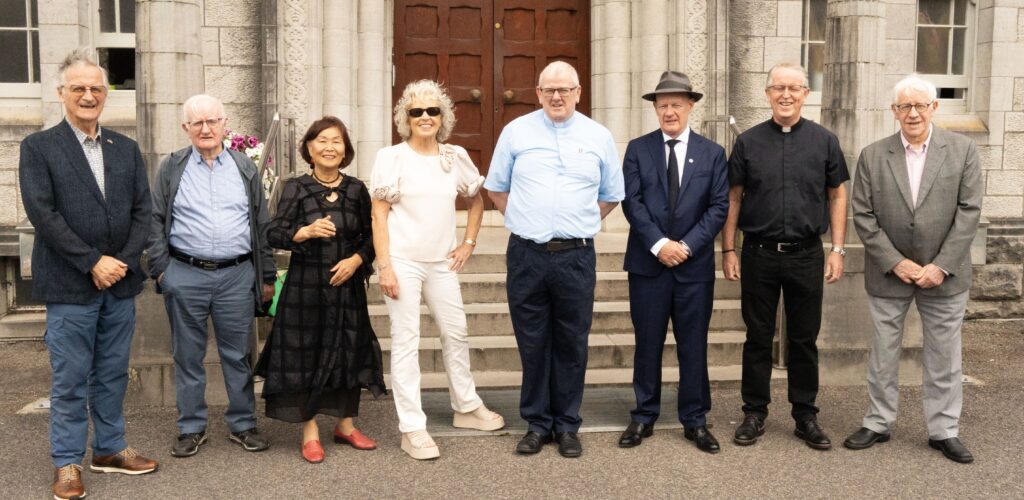 Eight adults standing in a row on stone steps in front of a large building, dressed in business and smart-casual attire.