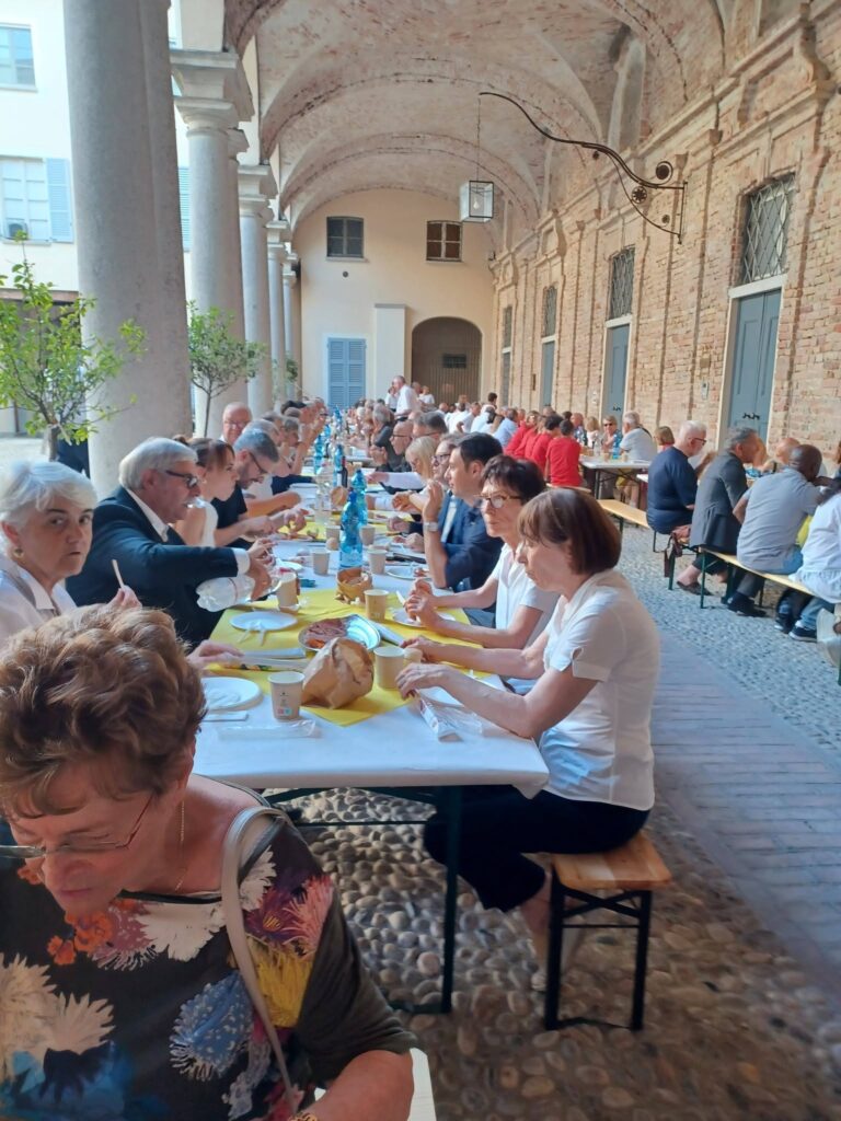 Long row of people dining at an outdoor courtyard banquet under brick arches, with yellow tablecloths and bread and bottles on the tables.