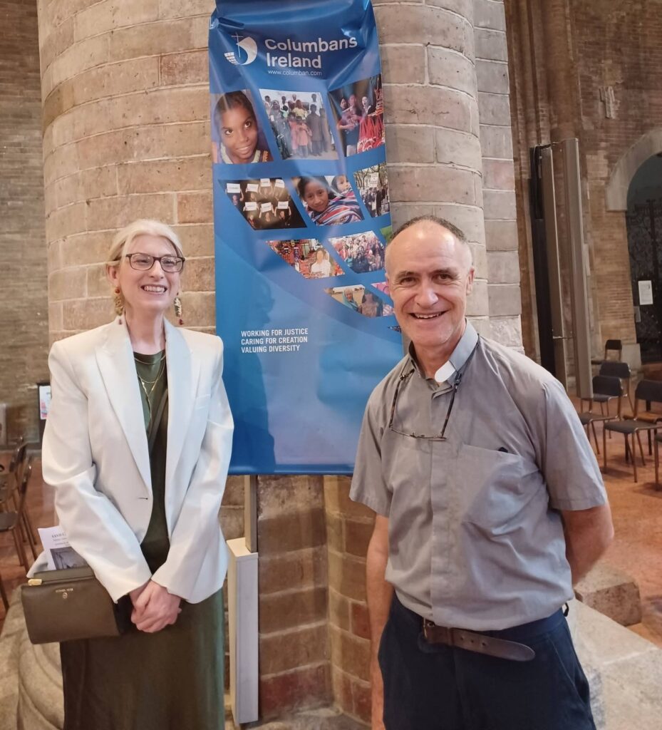 Two adults stand beside a blue Columbans Ireland banner in a stone-hall interior, smiling at the camera.