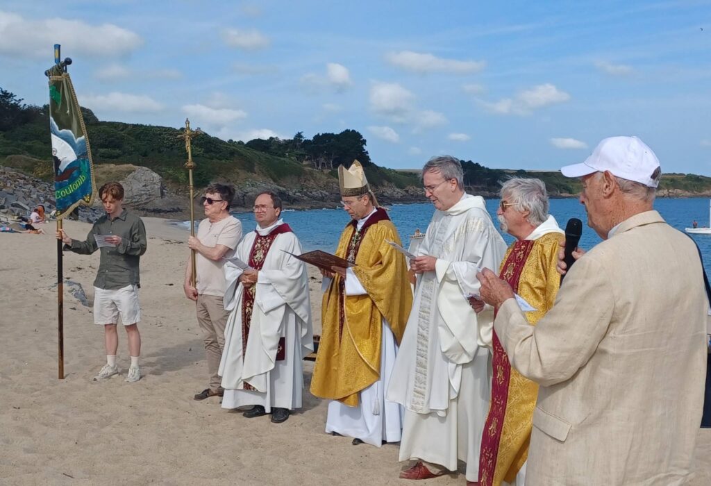 Clergy in ceremonial robes stand on a sandy beach during a coastal religious procession, with banners and a speaker.