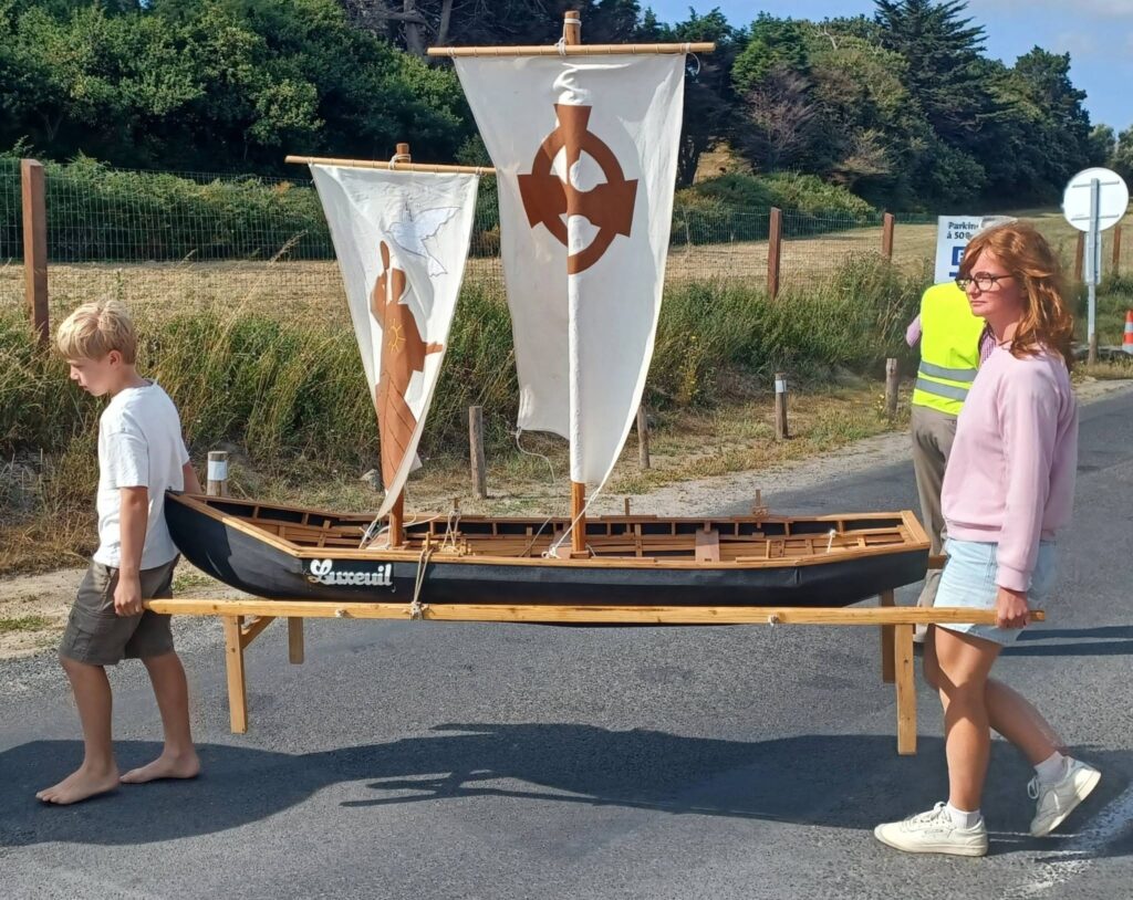 Two people, a boy and a woman, stand beside a small wooden sailboat on a wheeled trailer outdoors on a sunny day.