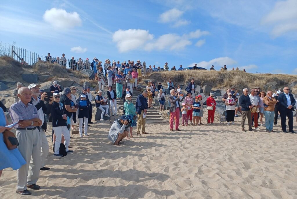 Large crowd of people gathered on a sandy beach with steps and a wooden fence under a blue sky with clouds.