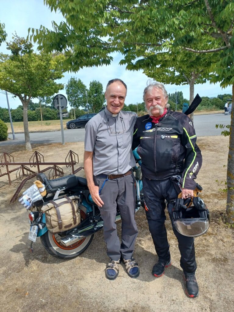 Two men in motorcycle gear posing beside a loaded touring motorcycle with panniers outdoors under trees on a sunny day, smiling at the camera.
