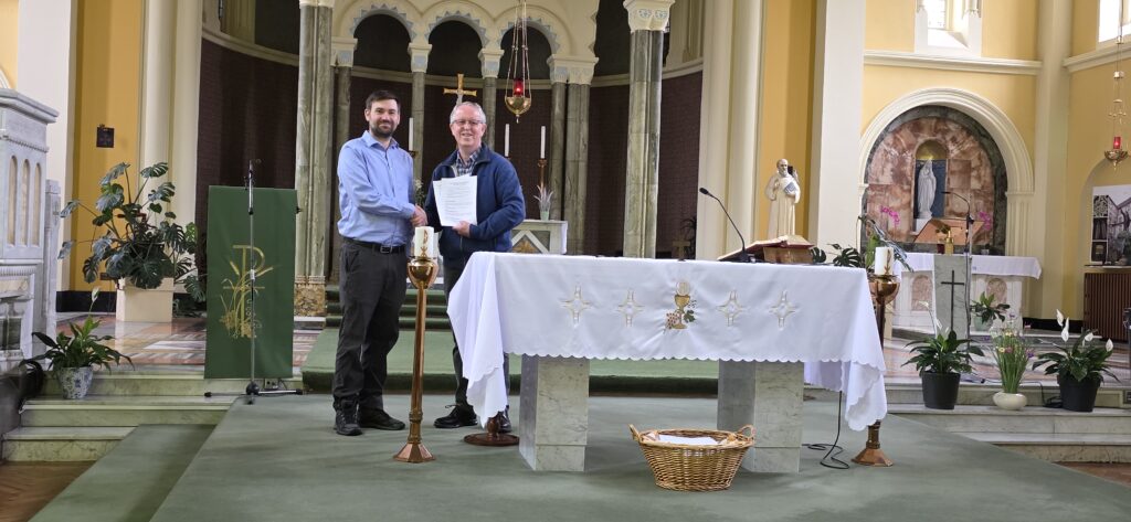 Two men shake hands at the church altar, one holding a document, with candles and marble columns behind them.