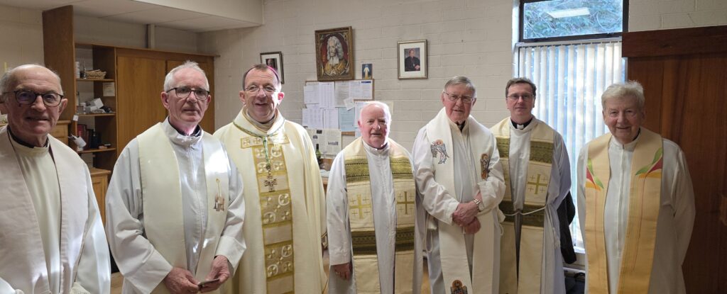 Seven priests in white liturgical vestments standing indoors, posing for a photo in a room with religious icons and a bulletin board walls behind them.