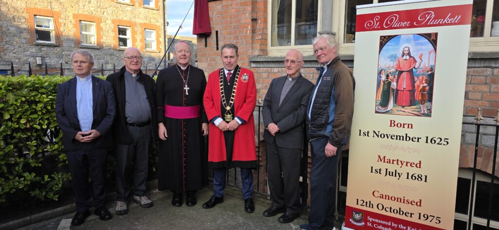 Six men, mostly clergy, stand outside a brick building next to a tall banner about St. Oliver Plunkett.