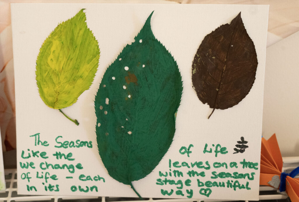 Canvas painting of three painted leaves (lime green, teal with white dots, dark brown) on white backdrop with green handwritten text about life and seasons.