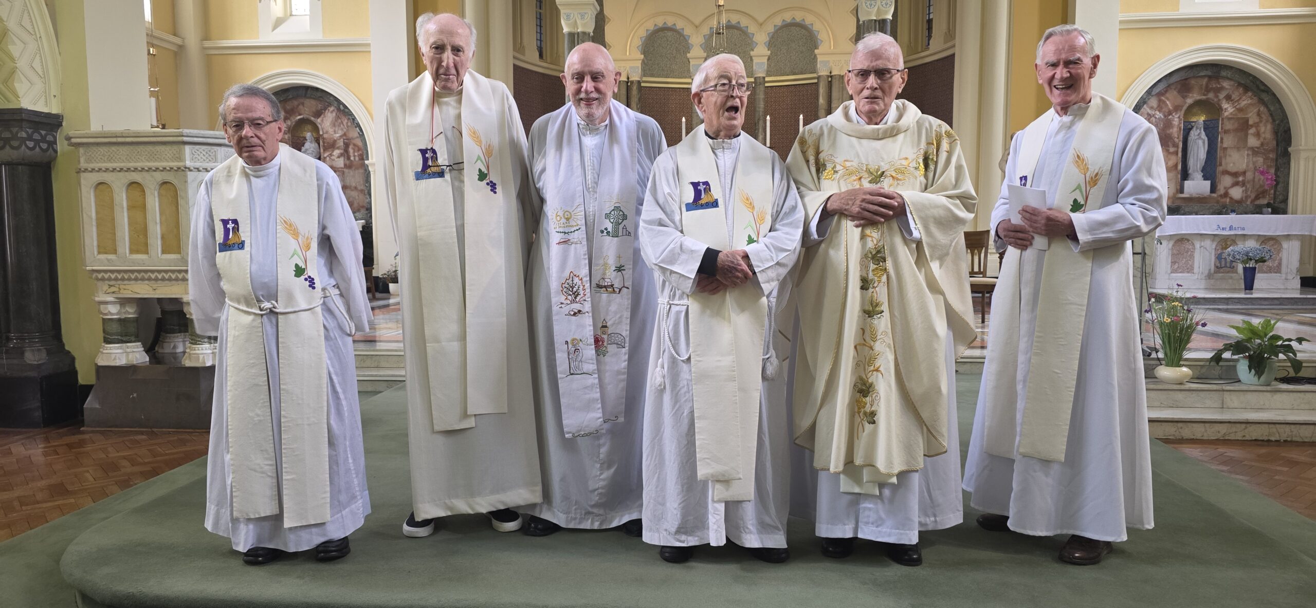 Seven Catholic priests stand together in white vestments at a church altar, smiling in a grand sanctuary with gold accents and arches.
