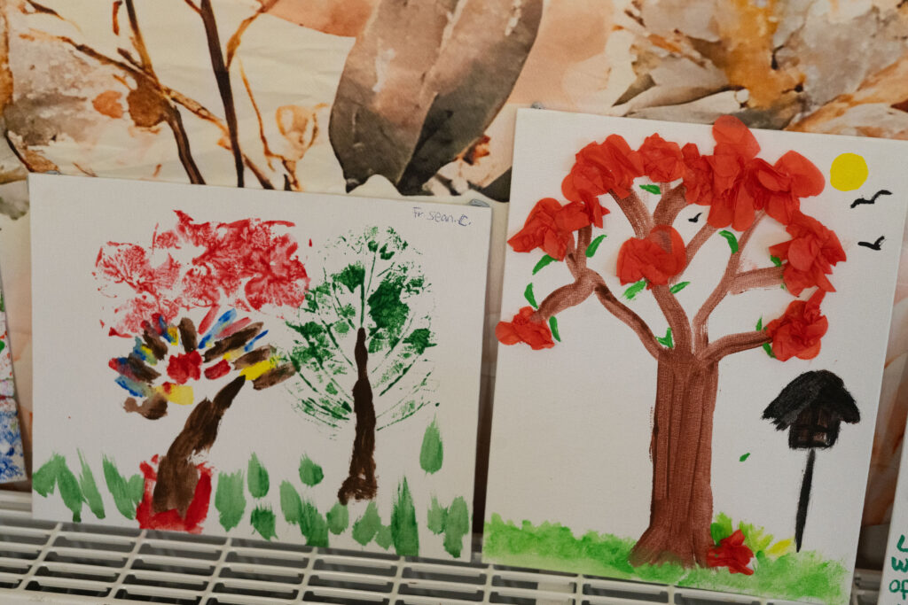 Child-made artwork featuring a tree with red paper leaves and a green grass border on white canvas