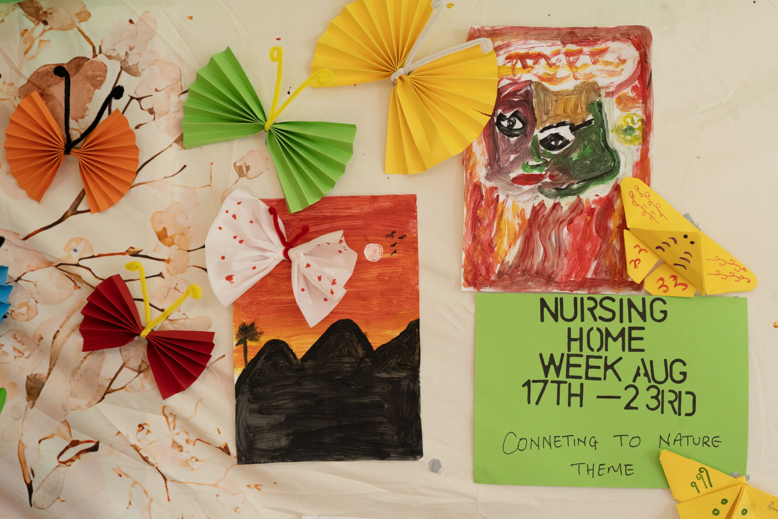 Colorful wall collage with folded paper fans in orange, green, and yellow, artwork on red and green backgrounds, and a green poster reading 'NURSING HOME WEEK AUG 17TH–23RD' with the tagline 'Connecting to nature' handwritten below.