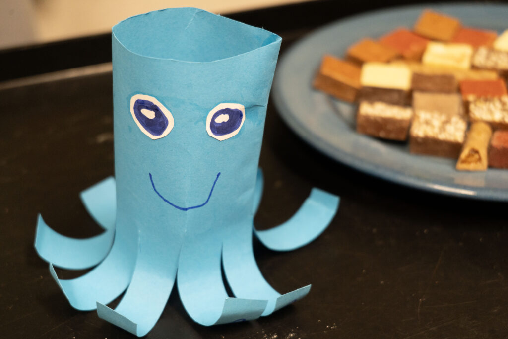 Blue paper origami octopus with drawn eyes and a smile sits on a dark surface, cookies on a plate visible in the background as a snack.