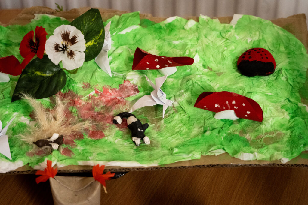 Colorful paper‑craft diorama of a green landscape with red mushrooms, origami cranes, and small figures.