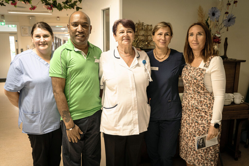 Five healthcare workers stand side by side in a bright hallway, smiling at the camera. From left: a nurse in blue scrubs, a man in a green polo, a nurse in a white coat, a staff member in navy scrubs, and a woman in a floral dress.