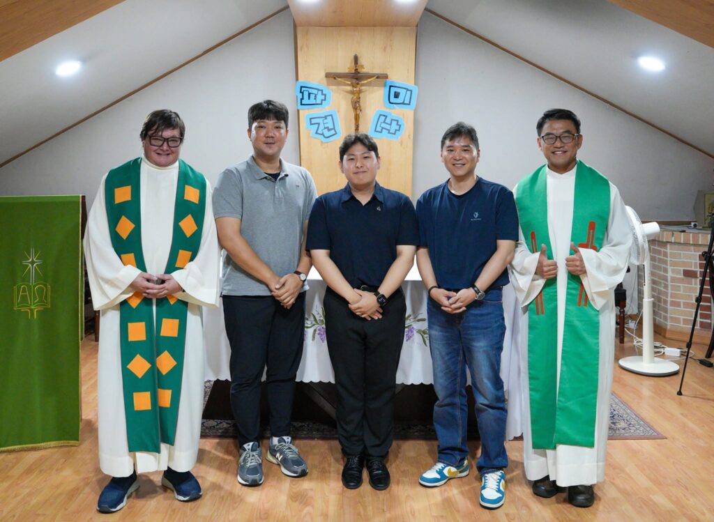 Five men pose for a group photo in a church, two priests in white robes with green stoles flanking three men, crucifix backdrop.